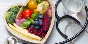 A bowl of fruit and vegetables in a heart shaped bowl with a stethoscope and glass of water beside it.